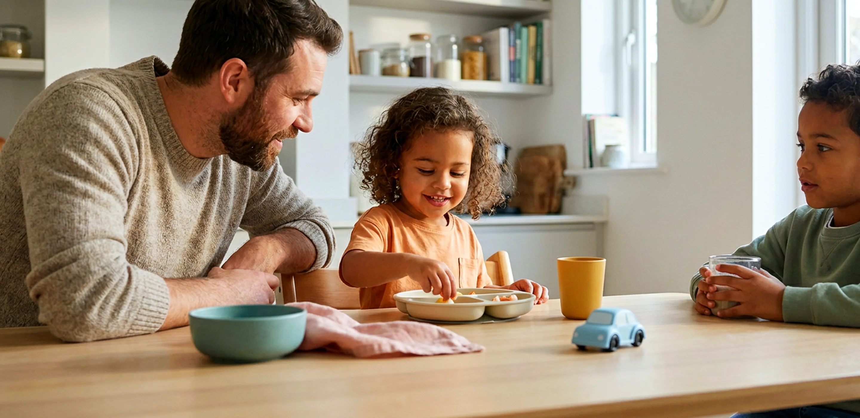 A father leaning in beside his toddler daughter in a high chair as she picks food off her plate, her older brother sitting further along the same kitchen table